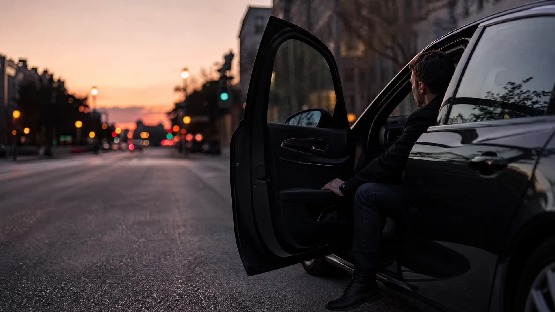 A man in dark clothing sits in the open rear doorway of a dark car parked on a city street at dusk, with blurred streetlights and traffic lights in the background.